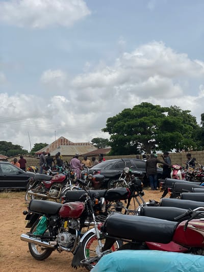 The image depicts an outdoor scene in the city of Kassa, Nigeria, with a substantial gathering of people and numerous motorcycles. The ground is covered in dirt. In the background, there are buildings with corrugated metal roofs, a large, leafy tree, and a stone wall. The sky is overcast with scattered clouds.

A group of men are seen standing and interacting among themselves, some gathered around motorcycles and cars. Several black and maroon motorcycles are prominently parked in the foreground and middle ground, some with riders or passengers seated. Two black cars are also visible. The overall impression is of a community gathering, possibly related to transportation or a local event.