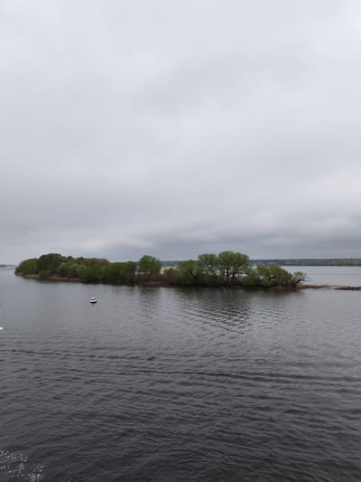 The image depicts an island covered in trees in the middle of a body of water, likely a river or lake. The sky is overcast. A white buoy is visible in the water near the island. In the background, a distant shoreline with trees can be seen. The water is dark and rippled. The location is not explicitly stated in the image or provided in the given context. Therefore, it is not possible to confirm that the location is Sloboda, Russia without additional information.