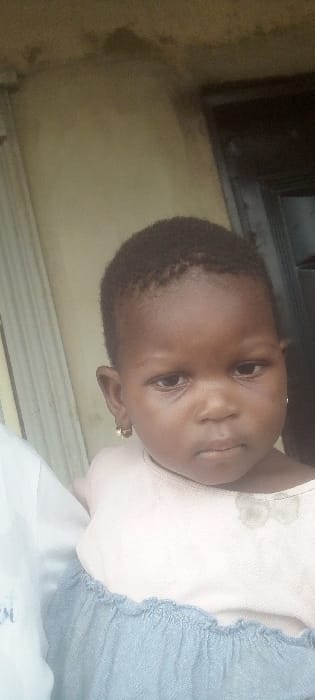 A close-up vertical portrait depicts a young child with dark skin and short, dark, curly hair, looking directly at the camera with a neutral expression. The child wears a light-colored, possibly pink, short-sleeved top with a faded, light-colored bow design on the upper chest, paired with a light blue, gathered lower garment. Small gold-colored hoop earrings are visible in both ears. On the left, a portion of another person, wearing a white garment with a dark blue or black pattern, is visible, suggesting the child is being held or standing adjacent to this individual. The background consists of a light brown wall exhibiting discoloration and staining, particularly in the upper half. To the right of the child, a dark brown or black vertical structure, consistent with a door or door frame, is present. A vertically striped light-colored element is visible on the far left. The scene is located in Agunmo, Nigeria.