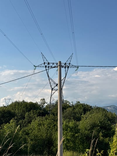 In the city of Dubovik, Montenegro, a power line pylon stands tall against a bright blue sky filled with scattered clouds. The pylon, made of concrete and steel lattice, supports thick electrical cables that stretch across the frame. Behind it, a dense forest of lush green trees covers the landscape, leading up to distant, hazy mountains. The scene captures the juxtaposition of industrial infrastructure with the natural beauty of the Montenegrin countryside.