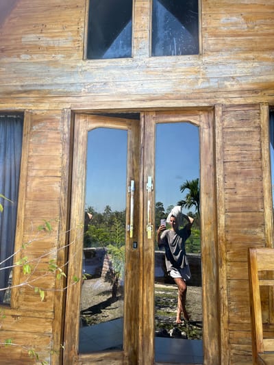 A full-body reflection shows a person standing in front of a wooden structure with glass doors. The person is holding a white towel on their head and taking a selfie with a smartphone. They are wearing a grey t-shirt and grey shorts. The reflection also shows a lush green outdoor environment with palm trees and a blue sky, indicative of a tropical location like Tetebatu, Indonesia. The wooden exterior of the building appears weathered, with visible planks and a rustic finish. A chair made of similar wood is partially visible to the right of the doors.