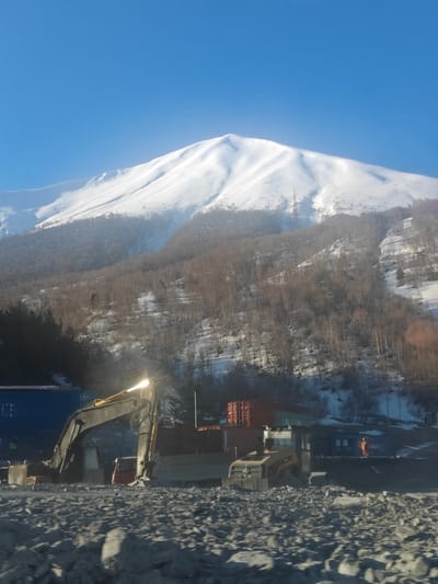 This image captures a winter landscape in Almasiani, Georgia, featuring a majestic snow-capped mountain presiding over a construction site.
The upper portion of the frame is dominated by a clear, bright blue sky. Below it, a large mountain peak is entirely covered in pristine white snow, displaying soft undulations and ridges. As the slope descends, the snow gives way to a dense forest of leafless deciduous trees, indicative of winter or early spring, with patches of snow still clinging to the ground between the trees. Electrical pylons are visible on the mountainside.
In the mid-ground, a scene of industrial activity unfolds. A large, dark-colored excavator with a prominent yellow boom is positioned on the left, its working light casting a bright glow. To its right, another piece of yellow construction equipment, possibly a roller, is visible. Several industrial containers are scattered around the site; a large blue container is on the far left, and multiple red/brown containers are positioned further right, suggesting temporary storage or offices. A small red vehicle is partially obscured behind the excavator.
The foreground consists of rough, rocky terrain, consistent with a construction or quarry site. On the far right, a worker dressed in bright orange safety gear stands near the containers, observing the scene.
The overall impression is one of active work taking place in a cold, mountainous environment under a clear, sunny sky, with strong sunlight creating contrast and reflections. There is no visible text.