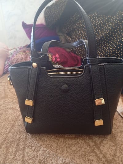 A black, textured leather handbag with gold-toned hardware is displayed in a close-up, eye-level shot. The bag has two top handles and a zippered top closure with a gold zipper. Decorative straps with gold buckles are attached to the front of the bag. The handbag is placed on a beige, carpeted surface. In the background, colorful fabrics are visible, including a patterned fabric with gold speckles and a purple fabric. The overall setting is consistent with an indoor environment, potentially a home. No specific landmarks or features place this image within the city of Hisor, Tajikistan.