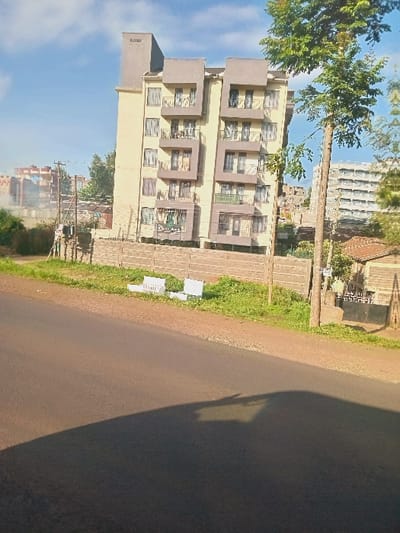 The image, when correctly oriented, depicts an outdoor scene in Wangige, Kenya, likely captured from a vehicle or roadside.

**Subjects:** The primary subject is a multi-story building, appearing to be an apartment complex, with a light yellow facade and distinctive greyish-brown framed balconies or window sections. A tall, leafy tree stands to the left of this building, contrasting with the bright blue sky. Further in the background, other buildings are partially visible. In the foreground, a paved road occupies the right side of the frame, bordered by a dirt shoulder and green grass on the left. A wooden utility pole stands along the roadside, in front of the main building. Several white, rectangular objects are placed on the ground near the grassy area. Shadows from an unseen object or vehicle are prominent on the road.

**Activity or event taking place:** The scene is static, with no visible human activity or events unfolding.

**Notable details:** The long, distinct shadows cast on the road suggest either early morning or late afternoon, indicating a low sun angle. The sky is clear and blue, with a few faint clouds, signifying sunny weather. The presence of multi-story residential buildings and a paved road points to an urban or semi-urban setting.

**Visible text:** On the upper-left side of the main building, the word "MSONGO" is clearly visible, written vertically. Towards the lower-right section of the building, on a white sign positioned near ground level, there is some blurry black text, which is difficult to decipher but may indicate property information or advertisements.
