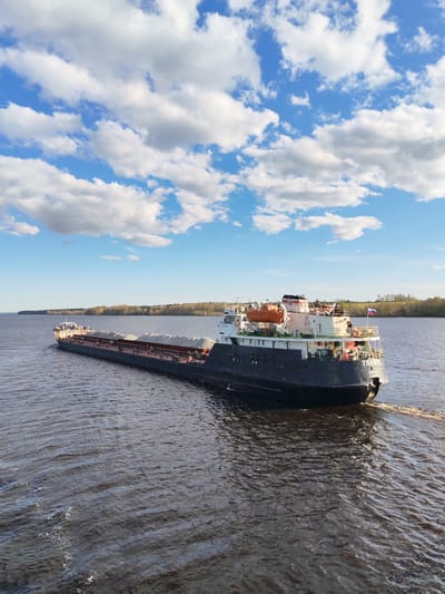 A barge, named "Tolvuya," is on a river in Yakunina Gora, Russia. The barge is black and carries a cargo of gravel or a similar material. The stern of the barge is visible, and it has a Russian flag. The river is brown and slightly rippled. In the background, there is a distant shoreline with trees. The sky is blue with scattered white clouds. The weather appears sunny and calm.