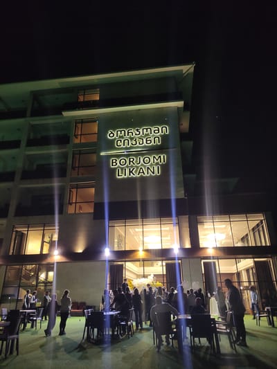 The image depicts the exterior of a modern building at night, illuminated by exterior lights and the interior lights of the building. The building features a facade with multiple floors, large windows, and balconies. A prominent illuminated sign on the building reads "BORJOMI LIKANI" in English, with Georgian script above it.
In front of the building, a group of people are gathered outdoors on a green surface that resembles artificial turf. There are several tables and chairs set up, suggesting an outdoor seating or event area. Some individuals are standing and conversing, while others appear to be seated. In the background, inside the building, the bright interior lighting illuminates the space, and a staircase is visible. White and yellow balloons are also present, indicating a celebratory occasion. The overall scene suggests an event or gathering taking place at a hotel or resort named Borjomi Likani in Borjomi, Georgia.