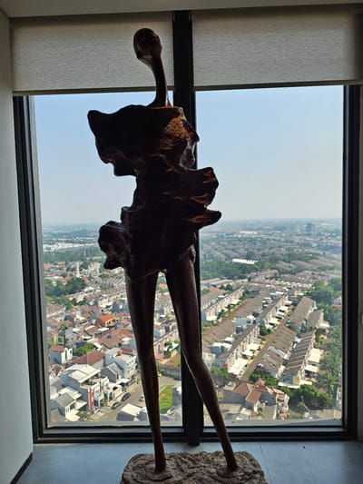 A full-body, dark brown, abstract sculpture of a standing figure with long, slender legs and an abstract head and torso is positioned in the foreground. The sculpture is silhouetted against a large window that offers a panoramic view of a densely populated urban landscape in Medan, Indonesia. The city below features a grid-like arrangement of houses with red and grey tiled roofs, interspersed with greenery. In the distance, taller buildings are visible, suggesting a sprawling urban environment. The sky is a pale blue, indicating a clear day. A light-colored roller blind is partially covering the top of the window. The lighting suggests it is daytime.