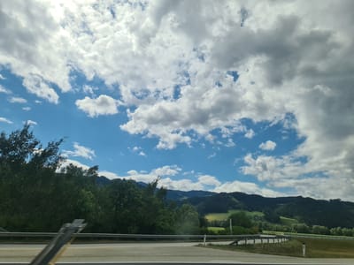 The image captures a roadside view, likely from a vehicle, on a partially cloudy day. The foreground shows a highway barrier and road markings, suggesting a driving environment. Beyond the barrier, a dense line of trees borders a landscape of rolling green hills and mountains in the distance. The sky is a mix of bright blue patches and large, white and grey cumulus clouds. The scene appears to be in a rural or semi-rural area, characterized by lush vegetation and natural terrain. The provided location, Sankt Marein-Feistritz, Austria, places this scene within a mountainous region known for its scenic beauty. There are no people or identifiable structures visible in the image, other than road infrastructure.