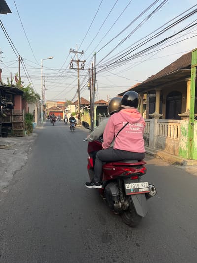 In Sugih Waras, Indonesia, two individuals are riding a red motorcycle on an asphalt road, heading away from the camera. The rider in the back, wearing a pink hoodie with the text "EX MACHINA CANGGU" and "JL. BATU MEJAH NO. 9 CANGGU" on it, has a black helmet. The passenger in front wears a gold helmet. The motorcycle has a license plate that reads "W 2918 NFC" with a sticker indicating "01.29". The street is lined with buildings, including houses with tiled roofs and fences, some appearing weathered. Utility poles with numerous wires are visible overhead. Other motorcycles are also seen on the road in the distance, indicating a typical street scene. The lighting suggests it is either early morning or late afternoon.