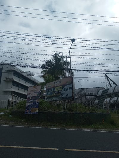 An outdoor scene shows a roadside view. A billboard advertises 'Rental' services with a picture of a van. A smaller banner displays travel destinations. Behind the billboard, there's a building with multiple windows, topped with a satellite dish. A palm tree is visible beside the billboard. Utility lines stretch across the sky above everything. A street lamp stands tall near the billboard. The foreground is a road with white lines.