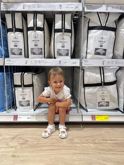 The image captures a happy young child lying playfully on a light-colored wooden floor in a retail store, likely a JYSK location in Ustroń, Poland. The child, with light hair and wearing a white top, pink bottoms, and white sandals adorned with a tiger-like design, smiles broadly at the camera. Their legs are bent at the knees, and hands are clasped, indicating a moment of candid joy during a shopping trip.
The background is dominated by white, packaged duvets or quilts neatly stacked on metal shelving units. Each package typically features the brand "KRONBORG" along with specific product lines such as "FJELLDAL", "PUTTEGGA", "BEERENBERG", and "SANDSTAD". The text "FIBRE DUVET" and "GOLD" are frequently visible, alongside descriptions like "EXTRA WARM" or "WARM" and various dimensions like "135x200 cm", "155x200 cm", and "200x220 cm".
Price tags in Polish złoty (zł) are displayed on the shelves. Examples include "1 SZT 150 zł" (1 piece 150 zł) and "1 SZT 300 zł" (1 piece 300 zł) for "PUTTEGGA" duvets, and "1 SZT 250 zł" (1 piece 250 zł) for a "FJELLDAL" duvet, noted with a yellow "PROMOCJA" (Promotion/Sale) tag. Another label reads "Im więcej, tym taniej" (The more, the cheaper), showing "2 SZT 469 zł" (2 pieces 469 zł) and "1 SZT 449 zł" (1 piece 449 zł) for a "SANDSTAD" duvet. Additional details on the packaging mention washing instructions (e.g., "Pranie: 60°C", "Pranie: 40°C"), material compositions ("Materiał poszycia: 100% bawełna", "100% poliester"), and descriptions like "Luksusowa kołdra" (Luxury duvet) and "Puch syntetyczny Trevira Fibelle" (synthetic down Trevira Fibelle). The indoor setting suggests daytime.