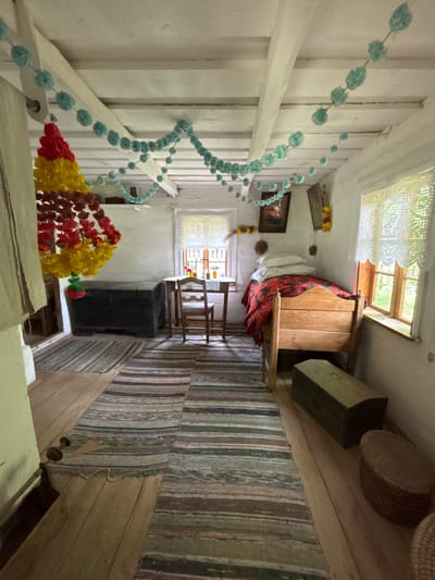 The image depicts the interior of a rustic room, likely a bedroom or living space, in a historical building in Sielachowskie, Poland. The room features whitewashed walls and wooden beamed ceilings. Decorations include garlands of blue pom-poms and a tiered, colorful floral arrangement hanging from the ceiling.

In the foreground, striped rag rugs are laid out on a wooden floor. To the left, a dark, large wooden chest is positioned against the wall, with a smaller, dark green chest further to the right. A woven basket is visible near the right wall.

Against the back wall, there is a simple wooden bed covered with a red and black patterned blanket and several pillows. Adjacent to the bed, to the right, is a wooden frame structure, possibly part of the bed frame itself.

A small wooden table with a chair sits in the center of the room, near a window. The window has white lace curtains. On the table, there are a few bottles or jars. Another smaller window is visible on the right wall, also with lace curtains.

On the walls, there are framed pictures and some hanging decorations, possibly dried flowers or foliage. The overall impression is one of a preserved historical dwelling, likely a museum exhibit or a traditional rural home. There are no people visible in the image.