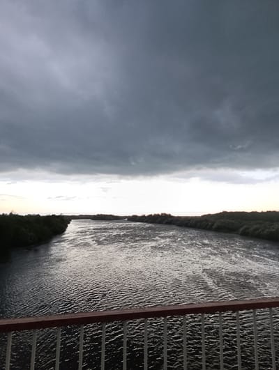The image depicts a river scene under a stormy sky. The river occupies the central and lower portions of the frame, its surface rippled by the wind and reflecting the dim light. Dense green foliage lines both banks of the river, creating a natural border. In the foreground, a railing with white vertical bars and a dark reddish-brown top rail suggests the viewpoint is from a bridge or elevated walkway. The sky is filled with heavy, dark gray clouds, indicating an overcast or impending storm. The light suggests it is either early morning or late evening, given the dim illumination and the muted colors. There is no visible text in the image. The overall mood is somewhat somber, dominated by the dramatic, cloudy sky and the vastness of the river. The location is identified as Naifel'd, Russia.