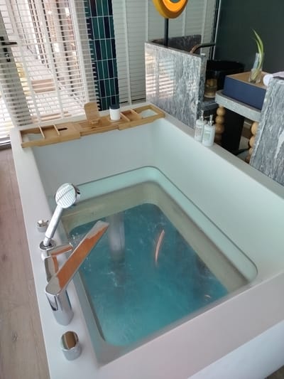 The image shows a close-up, high-angle view of a modern, minimalist bathroom with a bathtub filled with turquoise water. The bathtub is white and has a sleek, integrated design. A silver showerhead and faucet with a wooden accent are visible on the side of the tub. A wooden bath tray is placed across the bathtub's edge, holding a white cylindrical container and other small items.
In the background, to the left, are white horizontal blinds covering a window or door. To the right, a dark grey marble countertop is partially visible, with a black basin and a small glass vase containing a green plant. Two white pump bottles, presumably soap or lotion, are placed on a white surface near the marble countertop.
The overall impression is one of a luxurious and spa-like environment. The lighting is soft and diffused, suggesting an indoor setting. The scene appears to be in a hotel or high-end residence, likely in Kendhoo, Maldives, given the presence of the bath tray, often found in tropical resorts. There are no people or animals visible in the image. The floor is made of light-colored wooden planks.