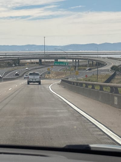 An eye-level, medium shot captures a scenic highway scene on a bright, overcast day in Vallejo, California, United States of America. The perspective is from the driver's seat of a vehicle, looking forward onto a multi-lane highway that curves to the right. The highway is well-maintained, with clear white lane markings.
In the foreground, the asphalt of the highway shows faint tire tracks. A concrete barrier with decorative openings runs along the right side of the road.
Further down the highway, a white pickup truck is traveling in the right lane, with its rear facing the viewer. To the left, other vehicles are visible in various lanes, including a dark-colored car and a white SUV.
The middle ground features an elevated highway interchange with curving ramps. A green highway sign indicates "Mare Island" with an arrow pointing to the right. Several yellow speed limit signs (30 mph) and traffic signs are also visible. The landscape beyond the highway includes scattered green vegetation and dry brush.
In the background, a body of water, likely a bay or strait, is visible, separating the highway from a distant mountain range. The mountains are hazy and appear to be a muted blue-gray. The sky above is a pale blue with scattered white clouds. The overall impression is one of a typical highway commute in a coastal California city.