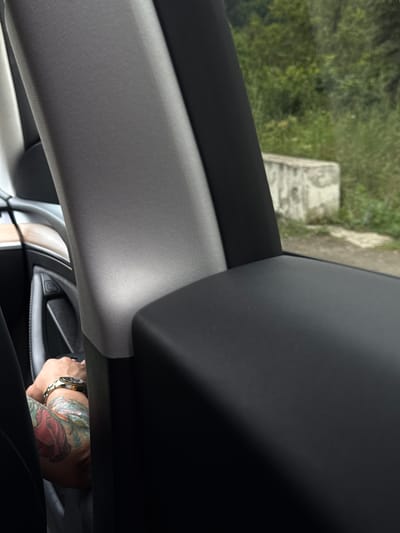 The image, taken in Vahagnadzor, Armenia, shows the interior of a vehicle. A person's arm, adorned with colorful tattoos of roses and feathers, is visible resting on a black surface, possibly an armrest or console. A gold watch with a black face is worn on the wrist. The interior of the car appears to be dark with fabric-covered pillars. Outside the vehicle, a blurred background of lush green trees and a concrete structure is visible.