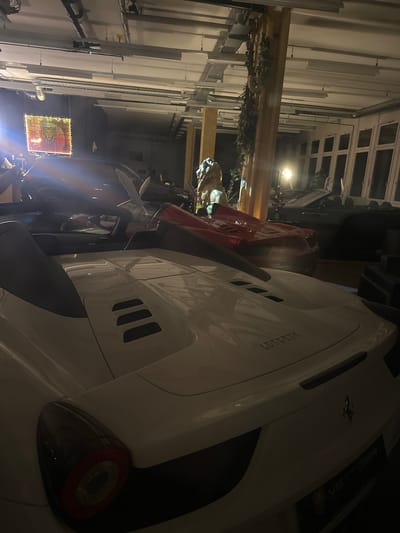 The image shows the interior of a garage located in Wetzikon (ZH), Switzerland. Several high-end cars are present, including a white Ferrari with the word "Ferrari" emblazoned on its side. The garage itself appears to be attached to a building or complex, evidenced by the presence of windows and a ceiling structure overhead. Other automobiles of different makes and models are visible, partially obscured by shadows and the main Ferrari in the foreground. In the background, a decorative sign or display can be seen illuminated with small lights. The overall environment suggests a private collection or storage facility for luxury vehicles.