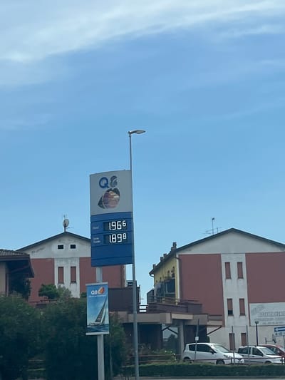 The image shows a gas station sign with prices for different types of fuel. The sign displays "Diesel" at 1.966 and "Senza Piombo" at 1.898, indicating the prices in euros per liter. Above these prices, the "Q8" logo is visible. To the left of the gas price sign, a smaller banner also features the "Q8" logo along with a sailboat. The gas station is situated in front of buildings that appear to be commercial or residential structures. The sky is clear and blue with some wispy clouds. Cars are visible parked in front of one of the buildings. The scene is captured on a sunny day. The location is identified as Castiglione delle Stiviere, Italy.