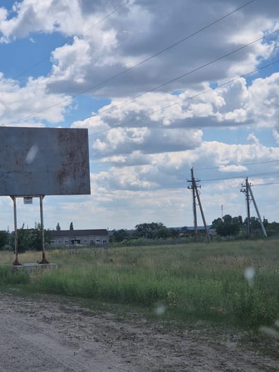 The image depicts an outdoor scene on a cloudy day. In the foreground, a dirt path runs horizontally. Behind the path, a grassy field extends into the middle ground. A large, weathered metal billboard stands on the left side of the frame, supported by metal posts anchored in a concrete base. A small sign is affixed to the left support post of the billboard. In the middle ground, a long, low-slung building with a dark roof and many windows is visible, surrounded by trees. Further back, two utility poles with wires strung between them are prominent. A more distant cellular tower can be seen behind the utility poles. The sky is filled with large, white, puffy clouds against a blue sky. The overall environment suggests a rural or semi-rural setting, possibly on the outskirts of a town. The location is identified as Pidhaitsi, Ukraine.