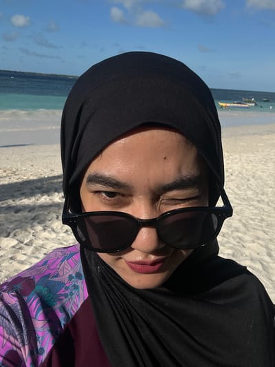 A woman wearing a black hijab and sunglasses winks at the camera while on a sandy beach. She is wearing a floral-patterned top. The ocean and sky are visible in the background, with a few boats in the distance. The location is identified as the city of A R A, Indonesia.