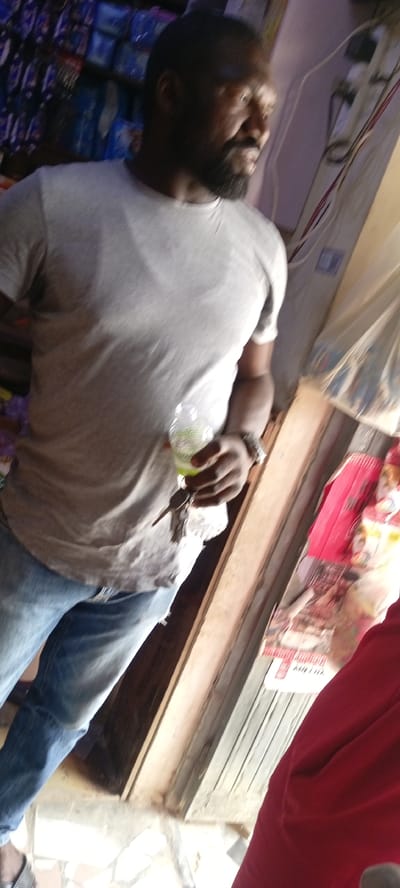 An indoor, slightly tilted medium shot captures a man standing in what appears to be a small shop or store in Gayawa, Nigeria. The man has a dark complexion, a beard, and short dark hair. He is wearing a light grey short-sleeved t-shirt, blue jeans, and a watch on his left wrist.

In his left hand, he holds a clear plastic bottle containing a bright green liquid, likely a beverage, along with a small bunch of keys. He is looking towards his right, out of the frame. His expression is calm.

The background is somewhat blurred but shows shelves stacked with various items, suggesting merchandise in a retail setting. On the far right side of the image, there is a wooden structure, possibly a doorframe or counter. Beyond this, a person in a red garment is partially visible. Further back, a red and white poster or packaging can be seen on a wall or surface, with the word "MILO" faintly visible in white text on a red background. The floor beneath him appears to be tiled. The lighting is bright, indicating it is likely daytime.