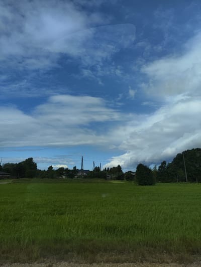 The image depicts a rural landscape in Ainaži, Latvia. The foreground is dominated by a lush green field of grass, with a visible gravelly path along the very bottom edge of the frame. In the middle ground, a line of trees separates the field from a distant village. Several buildings, including houses and what appear to be utility poles with wires, are visible amongst the trees. The background is filled with a dynamic sky featuring a mix of bright blue patches and large, fluffy white and grey clouds. The overall impression is that of a serene, overcast day in the Latvian countryside.