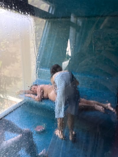 The image, shot through a rain-streaked window, shows a partial reflection of a person. The individual, wearing what appears to be a light-colored shirt and dark shorts, is partially obscured by the distorted reflection. The camera of the device taking the picture is visible in the upper left corner of the frame. The background shows some indistinct shapes and colors, suggesting an outdoor setting. There is no evidence of the city of Chornoye, Russia, in the image.
