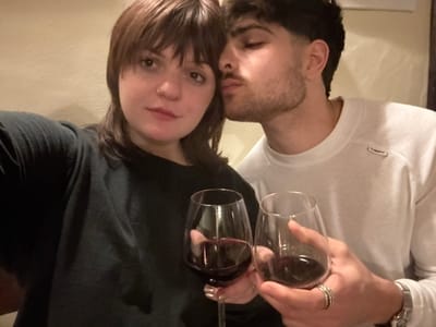 A man and woman pose with glasses of red wine. The man, on the left, wears a white sweatshirt and has multiple rings on his hand. The woman, on the right, has bangs and is wearing a dark top. They hold their glasses close to their faces, smiling at the camera. The image appears to be taken indoors, possibly in a home setting. The photo is rotated sideways.