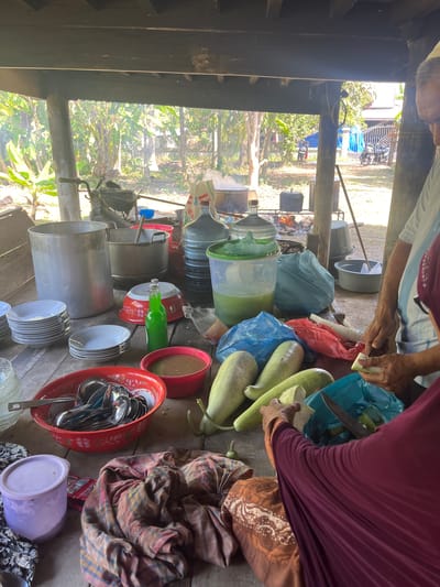 In Peurupok, Indonesia, a group of people are gathered around a rustic wooden table under a covered outdoor area, likely preparing a meal. The scene is dominated by cooking and food preparation items. Large metal pots, plastic water jugs, and stacks of plates are visible. Several people are actively involved in the preparation, with one person seen cutting vegetables and another seemingly preparing something in a red bowl. The environment appears to be outdoors, with lush greenery and trees visible in the background, suggesting a rural or semi-rural setting. The overall impression is one of community gathering and shared food preparation.