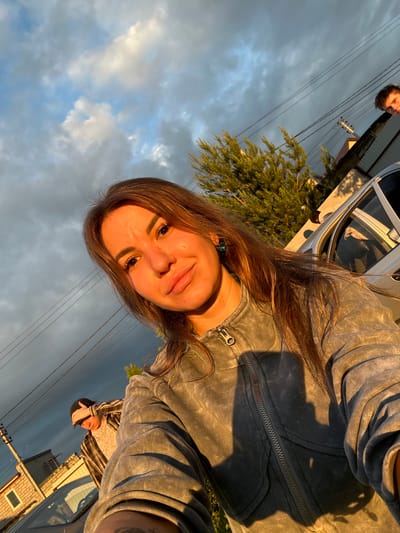 The image depicts an outdoor scene in Bol'shaya Mos', Russia, illuminated by golden hour sunlight. A young adult female, positioned in the foreground, is taking a selfie, looking directly into the camera. She has long brown hair and wears a grey zippered jacket with a prominent zipper pull and visible seams. A green stud earring is visible in her right ear. Her face is warmly lit on the right side by direct sunlight, casting shadows on the left. In the background, to the right of the woman, a silver-colored car with an open passenger door is visible. A young adult male's head is seen peeking from behind this car, looking toward the camera. To the left, a second young adult male is partially visible behind what appears to be another car's windshield. He is wearing a dark cap and a striped shirt, with his right hand raised toward his forehead. The background includes a dense green coniferous tree, utility poles, and multiple overhead power lines stretching across the frame. A light-colored brick building is partially visible on the far left. The sky is largely overcast with significant cloud cover, transitioning from lighter clouds on the left to darker, more ominous clouds on the right.