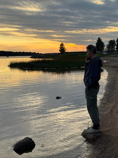The image depicts a man standing on a rock at the edge of a body of water, possibly a bay or lake, during sunset. The man is wearing a dark hoodie and green cargo pants, with his right hand near his face, suggesting he might be on a phone call. He is facing away from the camera, looking towards the horizon. The sky is filled with dramatic clouds, with streaks of orange and yellow from the setting sun illuminating the scene. The water is calm with visible ripples, reflecting the colors of the sky. A patch of tall grass and reeds is visible along the shoreline in the middle ground, and further in the distance, trees and buildings can be seen. The shoreline is sandy and appears to be a natural setting. The overall impression is one of quiet contemplation or a moment of connection amidst a natural landscape at dusk. While the prompt mentions the city of Glebychevo, Russia, this location is not explicitly discernible from visual cues within the image itself.