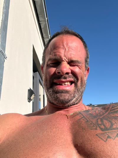 A low-angle, inverted close-up captures a shirtless man under a clear blue sky. The man, with graying dark hair and a beard, is positioned above the camera, leaning downwards. His face is contorted in a strong grimace or laugh, with his eyes squinted tightly and mouth open to reveal his teeth. His skin appears tanned and flushed, suggesting sun exposure.
A prominent, intricate black tattoo covers his right shoulder and upper arm, featuring geometric and abstract patterns. A thin gold chain is visible around his neck. In the background, behind the man's head and body, the bright blue sky dominates the upper portion of the frame. Below him, architectural elements of a building are visible: a light-colored wall or roof, a gray gutter, and what appears to be a skylight or window. A small, black pyramidal object, possibly a vent or cap, sits on the roof surface.
The image conveys a bright, sunny outdoor setting, likely during the daytime, given the strong illumination and clear sky. The man's intense facial expression suggests either amusement, discomfort from bright sunlight, or a reaction to something unseen below him. The perspective makes him appear upside down in the frame. There is no visible text. The setting is an outdoor location, possibly on a building's roof or upper level in Bredene, Belgium.