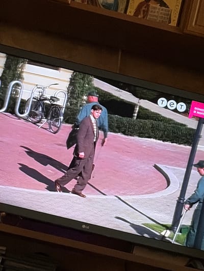 The image shows a screen displaying a scene with a man in a brown suit walking across a pink paved area. He is casting a long shadow. To his left, a bicycle is parked next to a silver rack. In the background, a person in a blue shirt and green hat is partially visible, and further to the right, another person wearing a green uniform and a cap is holding a white object. There are also decorative elements and logos visible on the screen, including "tet" and a pink sign with text in Cyrillic that appears to say "Закон жизни". The overall scene appears to be an outdoor setting, possibly in a park or public space. The location is identified as Osolynka, Ukraine.