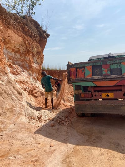 In Asaga Amangwu, Nigeria, a man in a green shirt and beige shorts is seen digging into a cliff face with a shovel. The cliff is composed of layered, reddish-brown and white soil. A large, colorful truck is parked near the man, partially obscuring the view. The sky is pale blue with scattered clouds. The ground around the truck is sandy and uneven.