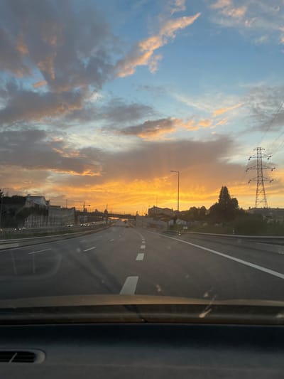 The image depicts a multi-lane asphalt highway at sunset or sunrise, viewed from inside a vehicle in Cidade da Maia, Portugal. The sky occupies the upper two-thirds of the frame, transitioning from clear blue at the top to vibrant orange and yellow hues at the horizon, with scattered clouds illuminated by the low sun.
In the immediate foreground, the dark dashboard of the vehicle is visible. The highway extends into the distance, featuring white dashed and solid lane markings. On the left side of the highway, a concrete retaining wall supports elevated terrain with various buildings. Distant vehicles are visible on the opposing carriageway.
On the right side of the highway, a vegetated area precedes residential structures and trees. A large, metal lattice-type electricity pylon stands prominently on the right, supporting multiple power lines that extend across the sky. Several tall streetlights line both sides of the highway, unlit.
An overpass or bridge structure crosses the highway in the mid-distance. No individuals or specific actions are discernible, only the environmental and infrastructural elements of the scene.