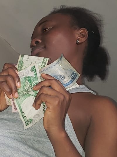 In Agunmo, Nigeria, a close-up, upward-angled shot captures an individual of African descent, facing partially right and looking upwards. The subject, wearing a light grey tank top and a silver hoop earring in their visible right ear, holds a quantity of Nigerian Naira banknotes with both hands close to their chest and chin. Visible denominations include ₦500 and ₦1000 notes. The money is crumpled and held loosely. The background is a plain, light grey surface.