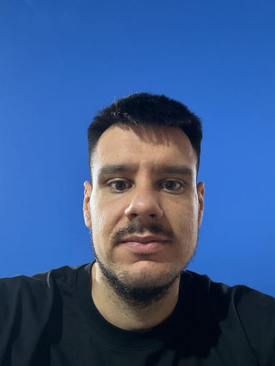 The image is a close-up, low-angle selfie of a man in Roquetas de Mar, Spain. The man has short, dark hair, a mustache, and stubble. He is wearing a black t-shirt. The background is a clear, vibrant blue sky. The lighting suggests it is daytime.