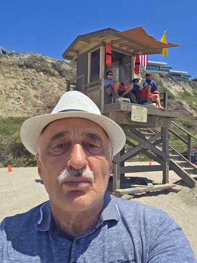 In this close-up, eye-level selfie, a man wearing a white fedora and a blue patterned polo shirt faces the camera. His gray mustache is prominent. Behind him, a wooden lifeguard tower stands on a sandy beach under a clear blue sky. Three individuals, presumably lifeguards, are seated on the platform of the tower. The tower is equipped with red rescue buoys and flags, including a yellow flag. The background features a steep, rocky cliff face and modern houses perched on the hilltop. A sign on the lifeguard tower reads "SAN CLEMENTE MARINE SAFETY" and lists various safety information. Orange traffic cones are visible on the sand.