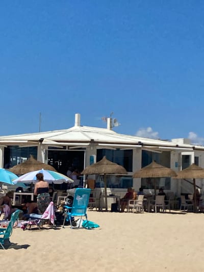 The image depicts a bright, sunny day at a beach in Conil De La Frontera, Spain. In the background, a white, modern beachside restaurant or cafe is visible with large windows and an outdoor seating area. Several thatched umbrellas and a colorful striped umbrella provide shade for patrons sitting at tables and chairs in front of the establishment. Beachgoers are scattered across the sandy shore, some seated under umbrellas, others standing or walking. Beach chairs, towels, and bags are present on the sand. The sky is clear and blue, indicating fair weather. The scene conveys a relaxed atmosphere characteristic of a popular beach destination.