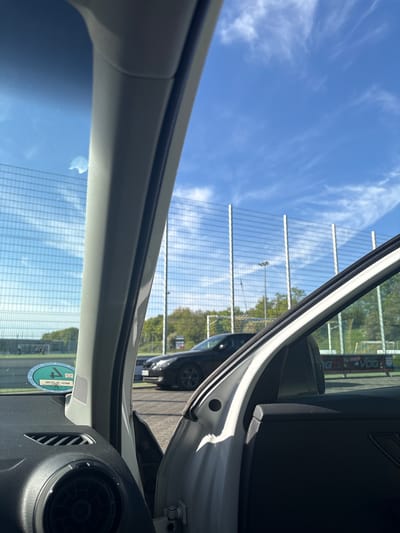The image, taken from inside a white car, shows the exterior of the vehicle through the open car door. A black car is visible outside. A large, tall metal grid-like structure stretches across the background, likely a sound barrier. The sky is blue with scattered clouds. A green circular sticker with white lettering is visible on the lower left portion of the windshield. Based on the vehicle license plate sticker, the location is Wassenberg, Germany.