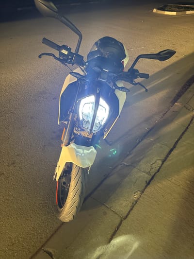 An overhead shot of a parked, white motorcycle, its headlight on, sits on an asphalt surface near a curb. The motorcycle appears to be a KTM Duke model, characterized by its angular design. Its handlebars are visible, and a helmet is positioned on the left side of the motorcycle. The background shows a concrete curb. There is no evidence to indicate that the image was taken in Mihailovo, Bulgaria.