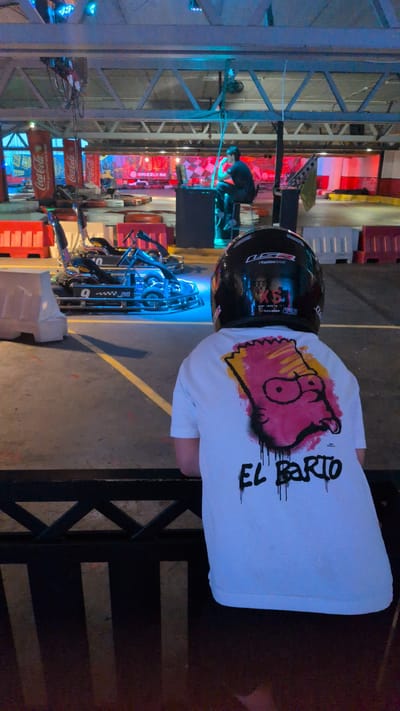 This media file shows a person wearing a black helmet and a white t-shirt with a graphic of Bart Simpson's face and the text "EL BARTO" on the back. The person is standing in an indoor go-karting arena, with go-karts visible in the foreground, some of which are lit with blue lights. The background features a checkered pattern, red and white barriers, and various advertisements, including several for Coca-Cola and one for GRIZZLY.SE. The lighting appears to be artificial, with a mix of colorful LEDs and ambient lighting, suggesting the event is taking place at night or in a dimly lit indoor space. The overall atmosphere seems to be one of leisure and entertainment, likely during a go-karting session or event. The location context indicates this is likely Östersund, Sweden.