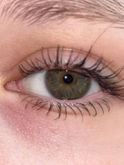 This is a close-up, macro photograph of a human eye. The iris is a striking hazel color with flecks of green and brown. The pupil is dark and reflects a small bright light source. The eyelashes are dark and appear to be coated with mascara, some clumped together. The skin around the eye is pale with visible pores and some subtle pink undertones. A fine network of lines, possibly representing wrinkles or the texture of the skin, is visible. The overall impression is of a well-lit, possibly indoor, environment. There is no visible text, indication of time of day, weather, or emotional state. The image is purely a detailed visual study of the eye itself.