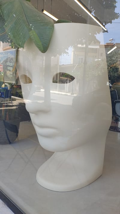 A white, oversized, stylized face sculpture is the dominant subject in this image, positioned in what appears to be a shop window or display area. The sculpture is smooth and featureless, except for two almond-shaped eye openings. Its base is rounded and rests on a light gray surface, likely the floor.
Reflections are prominent on the sculpture's surface, obscuring and merging with the actual environment. A large, green, tropical leaf from a plant, possibly a banana tree, is positioned to the left and slightly behind the sculpture's head, partially covering it.
Through the shop window, a street scene is visible, including buildings, trees, and the sky. The reflections also show elements of the interior, such as overhead fluorescent lights, a black salon chair with chrome accents, and what looks like a poster or signage on a wall to the far left. The signage on the left, partially obscured by reflections, displays text and an image of mountains, suggesting it might be a travel agency or a place with international themes.
The scene is likely located in Νέα Μηχανιώνα, Greece, given the context provided. The overall impression is one of a visually complex interplay between the sculpted form, the plant, and the mirrored world of the surrounding environment.