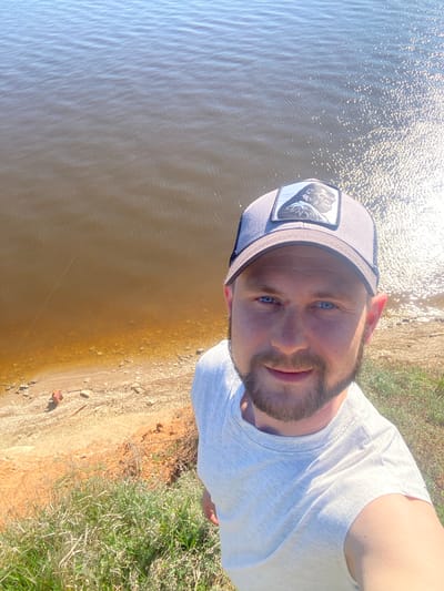 A selfie depicts a man in a gray cap with a gorilla patch and a white t-shirt, positioned near a body of water with ripples, and a shoreline transitioning from sand to grass. The location appears to be Шуран, Russia.
