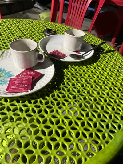 This media file features two white ceramic teacups, each resting on a saucer, positioned on a textured, bright green table. The table has a distinctive pattern of interconnected oval cutouts. Beside one teacup are two small, crimson red sachets of "GOLDEN CROWN" brand sauce. A silver spoon is visible on the saucer of the upper teacup. The scene appears to be outdoors, with hints of red plastic chairs visible in the background, suggesting a casual dining or cafe setting. The lighting suggests it might be daytime, with some shadows cast on the table. The visible text on the sauce sachets indicates they contain "SAUCE" and provide details about the manufacturer, "GOLDEN CROWN," and nutritional information, including "Net Wt. 5g." The setting is identified as Khangkor, India.