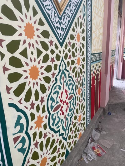 The image depicts a section of a building wall in Cutneuheun, Indonesia, adorned with intricate, multi-colored Islamic geometric and arabesque patterns. The primary wall features a cream background with a repeating pattern of interlocking dark green polygons, prominent orange eight-pointed stars, and smaller maroon six-pointed stars. A central vertical decorative panel showcases an elaborate, symmetrical curvilinear design in dark green, light blue, white, and maroon. To the right, a narrower vertical border consists of a cream section with dark green and white intertwined motifs, a light blue Greek key-style band, and vertical red and maroon arch-like elements. Adjacent to this, light peach-colored pillars and walls are visible in the background, displaying subtler cream geometric stenciling. A golden-colored trim is visible along the top edge of the patterned wall. The concrete ground in the foreground along the wall's base is unpaved and contains scattered debris, including white plastic bags and a red plastic object. No people, discernible actions, or interactions are visible.