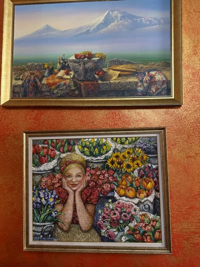 Two paintings hang on a textured orange wall. The top painting depicts a landscape with majestic snow-capped mountains in the background, a body of water, and a still life on a stone ledge in the foreground, featuring fruit, textiles, and a book. The bottom painting is a portrait of a woman with blonde hair, adorned with a floral headband, smiling with her hands supporting her chin. She is surrounded by an abundance of colorful flowers, arranged in bouquets and baskets. The lower left corner of this painting contains a handwritten inscription. The overall setting suggests an interior space, possibly a home or a gallery, located in Ozeretskoe, Russia.