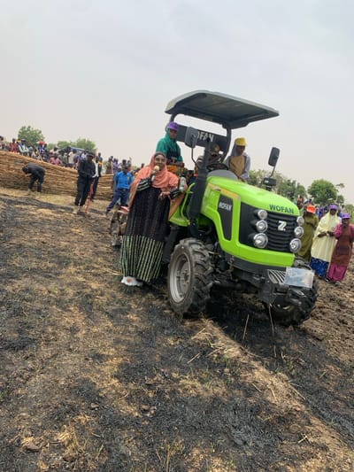 A woman in a black and white dress and headscarf is speaking into a microphone while standing on the ground next to a green tractor with the word "WOFAN" on the side. The tractor is parked in a field of burnt grass and has a canopy roof with three people sitting inside. There are several other people standing around the tractor. The location is Ringimawa, Nigeria, based on the coordinates 11.666 latitude and 8.415 longitude.