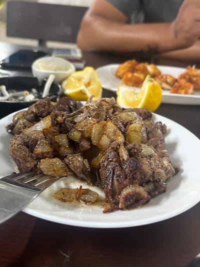 The image depicts an indoor dining scene in Karadjovo, Bulgaria. The primary subject is a white oval plate positioned centrally, filled with sautéed, dark brown meat pieces (consistent with liver) and caramelized golden-brown onion slices. A silver two-tined fork rests on the left side of the plate, partially submerged in the food. Liquid residue and small food particles are visible on the plate's surface. Two yellow lemon wedges are present in the mid-ground, one to the left of the main plate and another further right. In the out-of-focus upper left background, a black ashtray contains multiple white cigarette butts and ash, adjacent to a small white bowl, likely holding a condiment. Further to the blurred upper right, a second white oval plate displays several pieces of fried or breaded chicken. The table surface is dark brown. In the highly blurred background, a person's tanned forearms are visible, one featuring a faint tattoo, along with a portion of a grey short-sleeved garment. A dark rectangular object, possibly an electronic monitor, is discernible behind the person.
